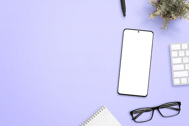 Smart phone with isolated screen for mockup on purple desk surrounded by glasses, pad, plant, keyboard and pen. Clean space beside for text