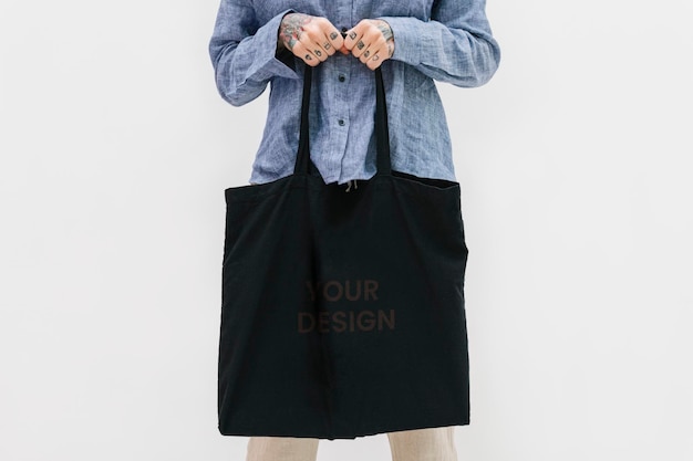 Tattooed woman in a blue linen shirt holding a black tote bag mockup