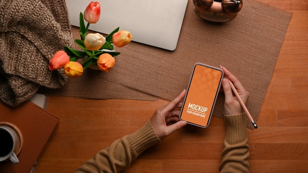 Top view of female hand using smartphone mockup with laptop and flower vase