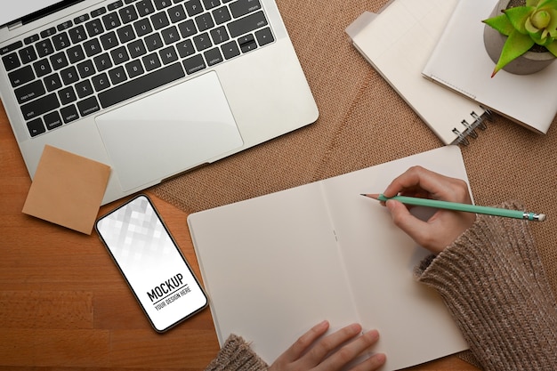 Top view of female hand writing on blank notebook on worktable with smartphone mockup