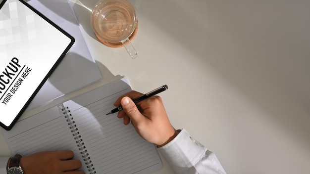 Top view of male hand writing on blank notebook while working with digital tablet mockup