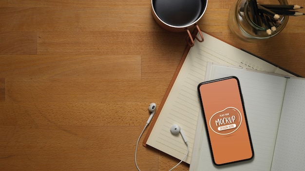 Top view of worktable with mock up smartphone on opened notebooks, earphone, mug, pencils