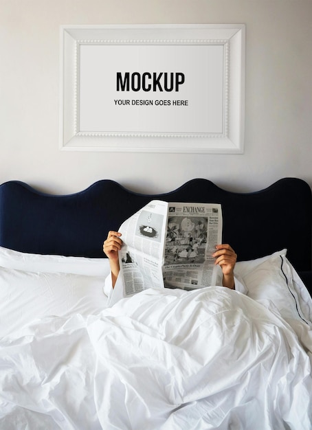 An unfeatured woman reading the newspaper in a bed with white sheets and a picture frame above