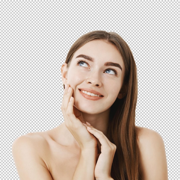 Vrouw die crème op haar gezicht aanbrengt, haar huid zacht en zacht voelt, droomend staat en verheugd met het resultaat, kijkt naar de linkerbovenhoek met een sensuele glimlach, raakt haar wang aan, poseert naakt over de grijze muur.