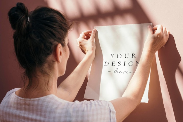 Vrouw die een wit postermodel vastmaakt aan een pastelroze muur