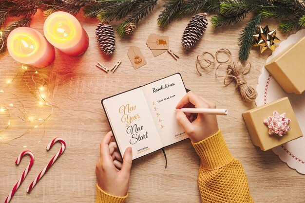 Vrouw hand schrijven in een nieuwjaarsresolutie notebook mockup met kerst decor op een houten tafel