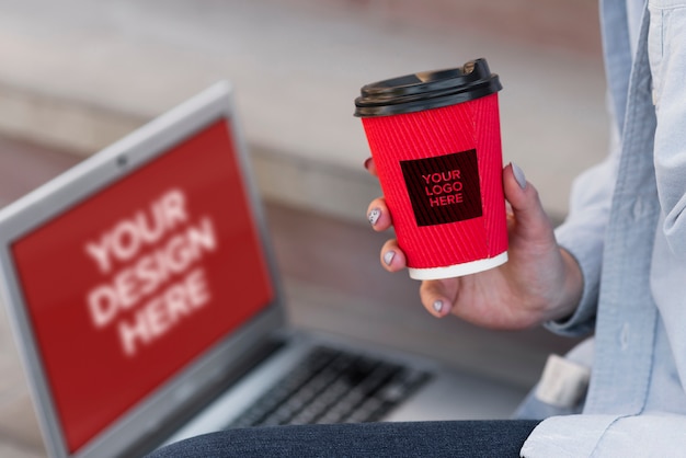 PSD woman holding a cup of coffee while sitting next to a laptop mock-up