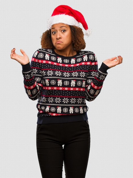 Молодая чернокожая женщина в модный рождественский свитер с печатными сомневающимися и пожимающими плечами плечами