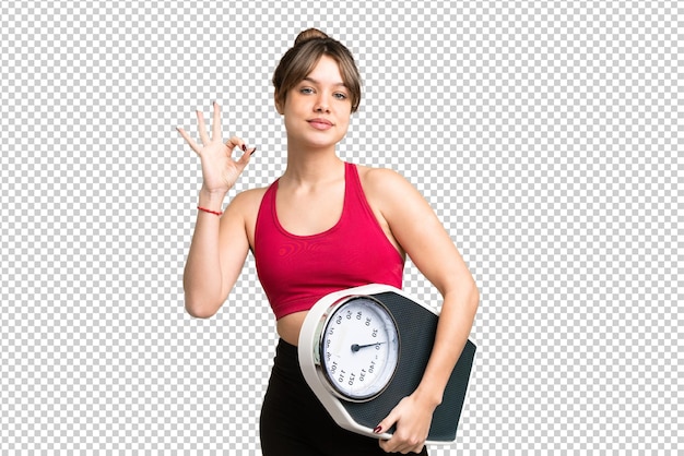 Young girl over isolated chroma key background holding a weighing machine and doing OK sign
