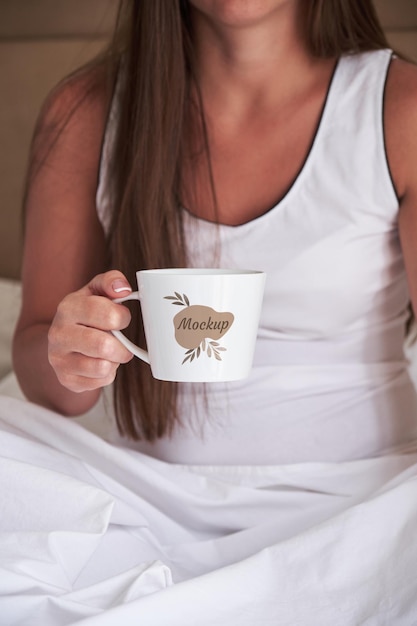 A young girl in a white tank top holds a white mug while sitting on the bed in the bedroom Mockup