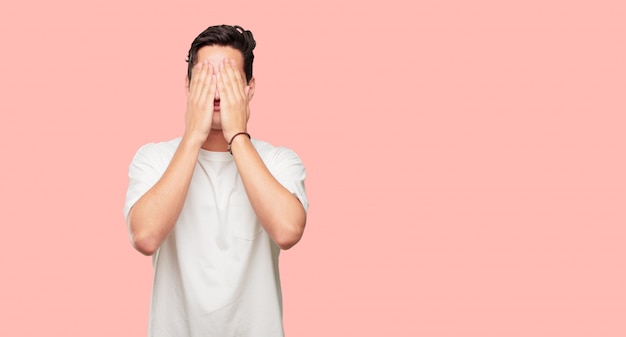 Young handsome man with a serious, scared, frightened expression, covering eyes with both hands