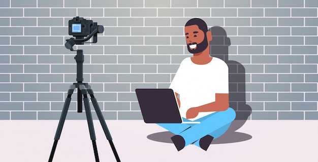 African american blogger using laptop recording video blog with digital camera on tripod live streaming social media blogging concept brick wall background full length horizontal