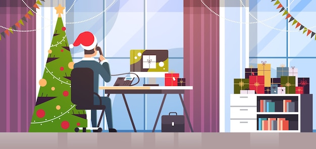 サンタクロースの帽子をかぶったビジネスマンが電話で話している職場で話しているビジネスマンギフトボックスでモニターを見て冬の休日のお祝いのコンセプトモダンなオフィスインテリア
