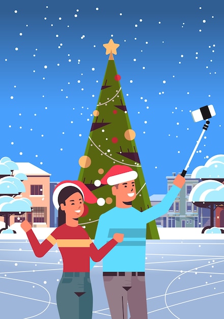 サンタの帽子をかぶったカップルがスマートフォンのカメラで自分撮り写真を撮るモミの木の近くに屋外に立っている男性女性メリークリスマス新年あけましておめでとうございます休日コンセプト都市景観背景縦図