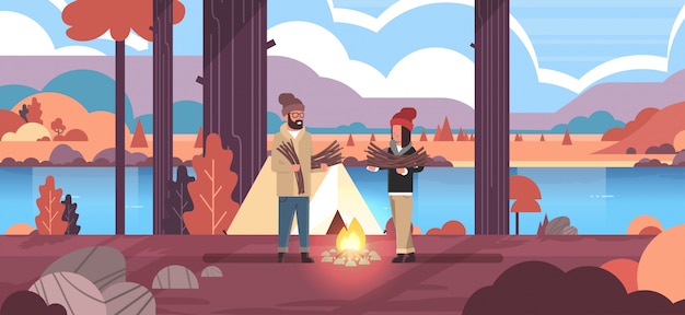 couple tourists hikers holding firewood man woman organizing fire near camp tent hiking camping concept autumn landscape nature river mountains 