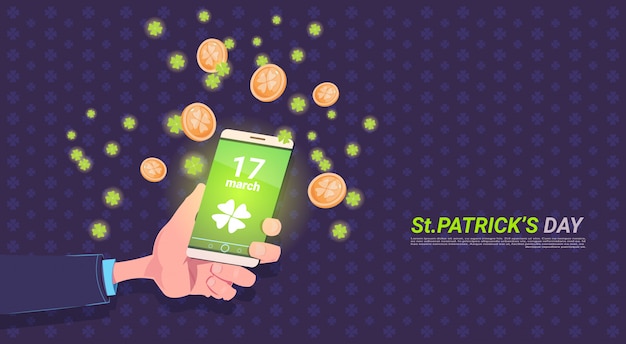 Smart phone della tenuta della mano con la foglia del trifoglio e le monete dorate sopra fondo felice del giorno di st patrick