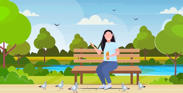 het brood van de vrouwenholding en het voeden troep van duif te zwaar meisje die houten bank zitten die het landschapsachtergrond hebben van het pret openlucht openbare park vlakke volledige horizontale lengte als achtergrond