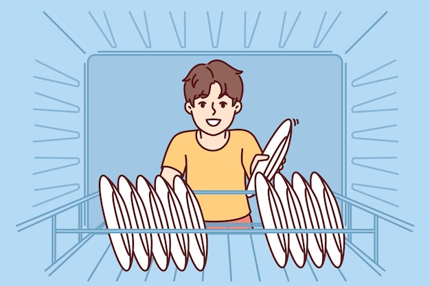 Маленький мальчик ставит посуду в посудомоечную машину, чтобы поддерживать гигиену и помогать родителям по дому Счастливый ребенок-подросток с удовольствием разгружает посудомоечную машину и смотрит на экран, улыбаясь