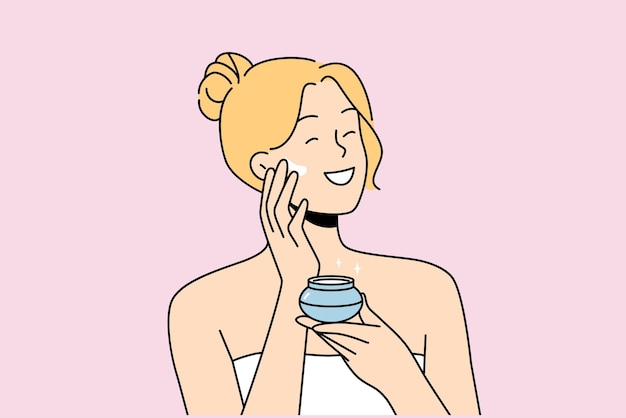 Woman applies antiaging cream to face to stay beautiful standing in bath towel after shower Happy blonde girl smiles and uses cosmetic cream to help prevent wrinkles and pimples