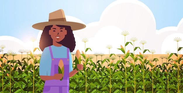 Contadina della tenuta della pannocchia della donna di campagna afroamericana in tuta che sta sull'orizzontale piano del ritratto di concetto di stagione di raccolta di agricoltura biologica del campo di grano