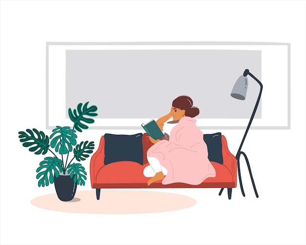 A young woman reads a book while sitting under a blanket on a red sofa in the apartment.