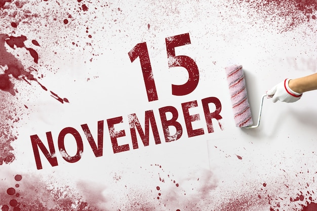 15 listopada. Dzień 15 miesiąca, data kalendarzowa. Ręka trzyma wałek z czerwoną farbą i pisze datę w kalendarzu na białym tle. Jesienny miesiąc, koncepcja dnia roku.
