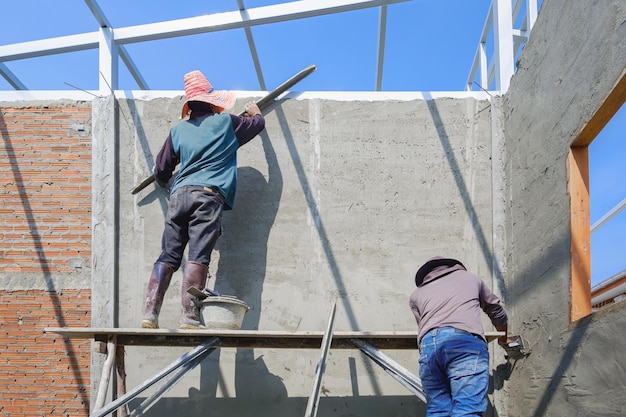 2 pracowników budowlanych na drewnianych rusztowaniach tynkuje betonową ścianę na budowie domu
