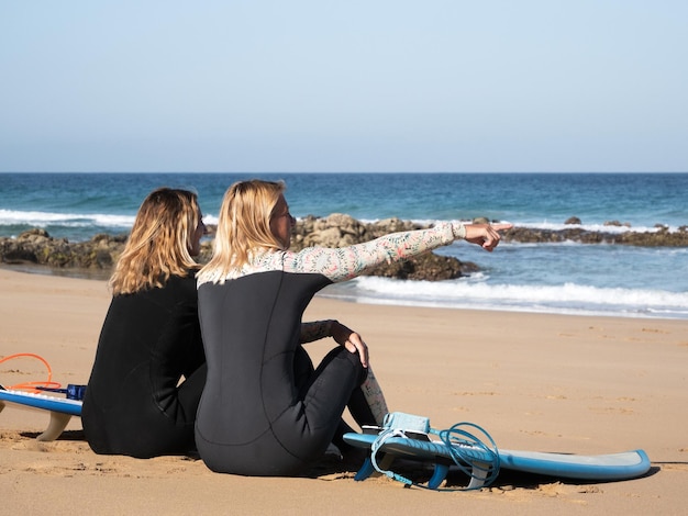 2 surferki siedzące na plaży obserwujące fale