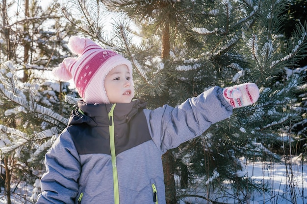 34-letnia dziewczynka w czapce i rękawiczkach w zimowym kombinezonie stoi na tle ośnieżonych sosen i jodły, odwracając wzrok