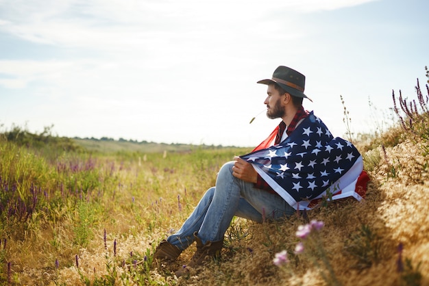 Zdjęcie 4 lipca. czwarty lipca. amerykański z flagą narodową. flaga ameryki. dzień niepodległości. patriotyczne wakacje. mężczyzna ma na sobie kapelusz, plecak, koszulę i dżinsy.