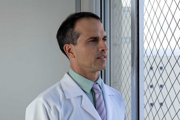 44-letni lekarz w białym fartuchu i krawacie patrzy w okno.
