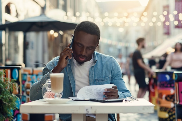 African American Biznesmen Czyta Dokumenty Papierowe I Pracuje Na Laptopie Siedząc Na Zewnątrz Kawiarni Z żarówkami.