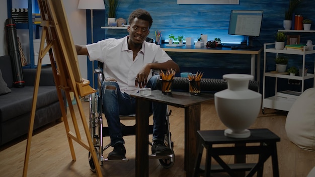 African american człowiek siedzący na wózku inwalidzkim podczas rysowania i projektowania arcydzieła wazy na stole. Czarny młody dorosły używający narzędzi, białego płótna na sztalugach i kolorowych ołówków