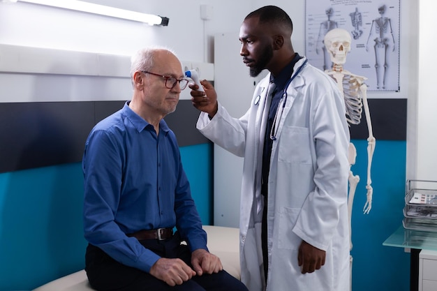 African american doctor doctor stawiając termometr na podczerwień na starszy mężczyzna czoło pacjenta pomiaru temperatury w biurze szpitala. Terapeuta wyjaśniający objawy grypy podczas konsultacji lekarskiej w