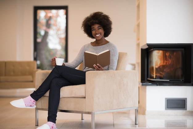 african american kobieta pije filiżankę kawy czytanie książki przy kominku. Młoda czarna dziewczyna z gorącym napojem relaksującym ogrzewaniem rozgrzewającym. jesień w domu.