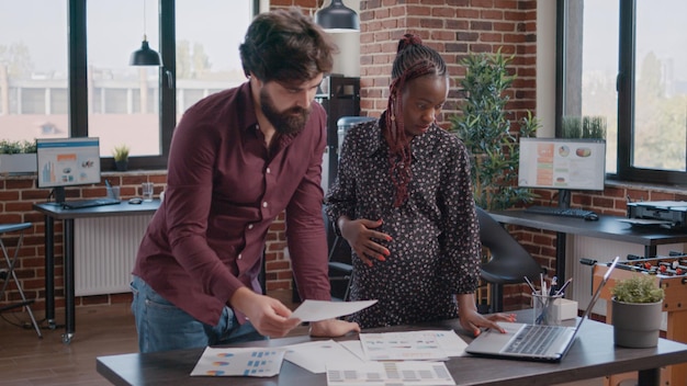 African American Kobieta W Ciąży Pracuje Nad Planowaniem Projektu Z Mężczyzną W Biurze. Ludzie Biznesu Robiący Pracę Zespołową Z Papierami, Rozmawiający O Rozwoju. Pracownica W Ciąży Spodziewa Się Dziecka