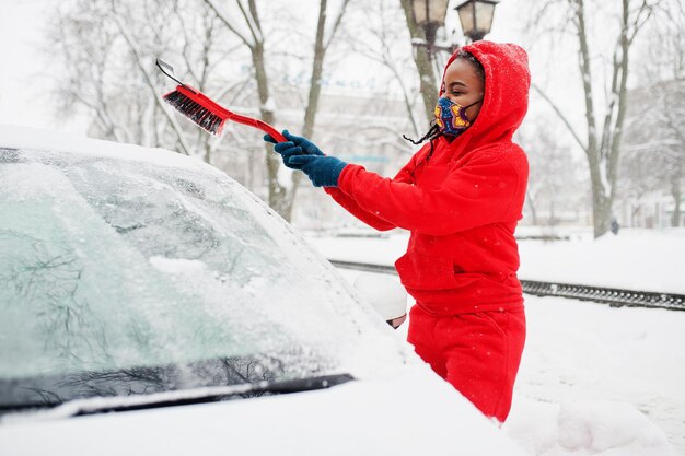 African american kobieta w czerwonej bluzie z kapturem i maską czysty samochód od śniegu w zimowy dzień.