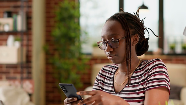 African American kobieta za pomocą smartfona do wiadomości tekstowych w sieci społecznościowej. Przerwa i rozmowa przez telefon komórkowy po pracy zdalnej na laptopie w domu, praca jako freelancer.