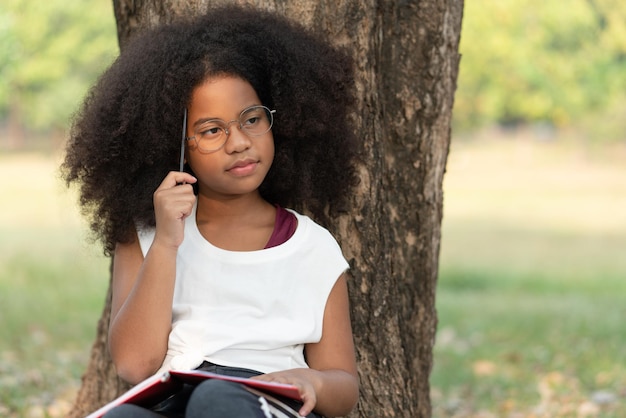 African American kręcone włosy młoda dziewczyna nosi okulary trzymać ołówek odrabiania lekcji pod drzewem w parku