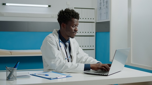 African american lekarz za pomocą laptopa siedząc przy biurku w sali szpitalnej. Czarny specjalista medyczny pracujący z technologią do konsultacji w nowoczesnym gabinecie w placówce medycznej