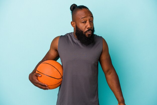 African american man grający w koszykówkę na białym tle na niebieskim tle wzrusza ramionami i otwiera oczy zdezorientowany.