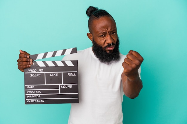 African american man trzymając clapperboard na białym tle na niebieskiej ścianie pokazując pięść do kamery, agresywny wyraz twarzy.