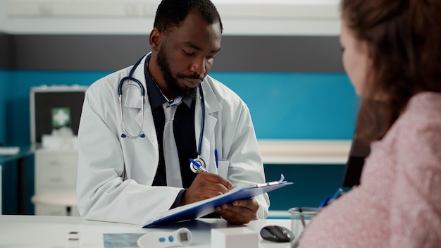 African American Medic Robienie Notatek Podczas Konsultacji Z Osobą W Ciąży, Pisanie Raportu Kontrolnego Na Dokumentach. Kobieta Z Ciążowym Brzuchem Prosi Położnika O Poradę Macierzyńską.