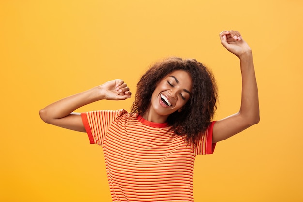 African American młoda kobieta w tshirt w paski, wyciągając ramiona, zamykając oczy z zachwytu i uśmiechając się na pomarańczowej ścianie