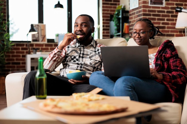 African american para jedzenie frytek i za pomocą laptopa przed telewizorem, oglądając film w programie kanału. Ciesz się rekreacją z komputerem i dostawą fast foodów na wynos.