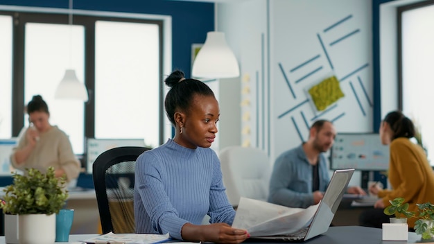 African american startup pracownik posiadający dokumenty biznesowe z wykresami, patrząc na ekran laptopa porównując dane. Kobieta w zajętym biurze analizuje statystyki marketingowe i roczne obroty.