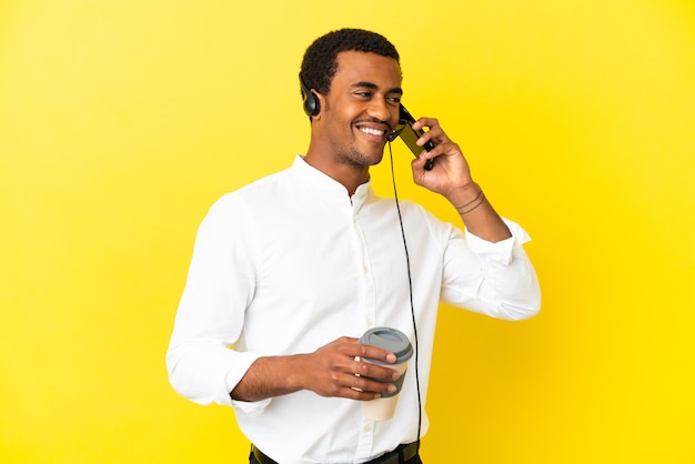 African American Telemarketer mężczyzna pracujący z zestawem słuchawkowym na odizolowanym żółtym tle, trzymający kawę na wynos i telefon komórkowy