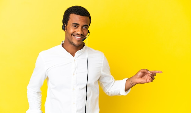 African American Telemarketer mężczyzna pracujący z zestawem słuchawkowym na odizolowanym żółtym tle wskazujący palec w bok