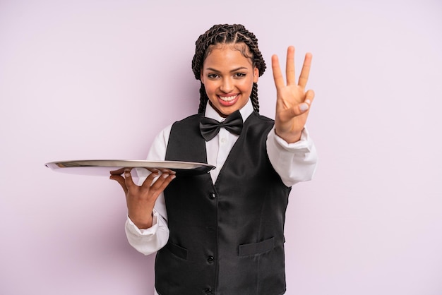Afro czarna kobieta uśmiechnięta i wyglądająca przyjaźnie, pokazująca kelnera numer trzy z tacą