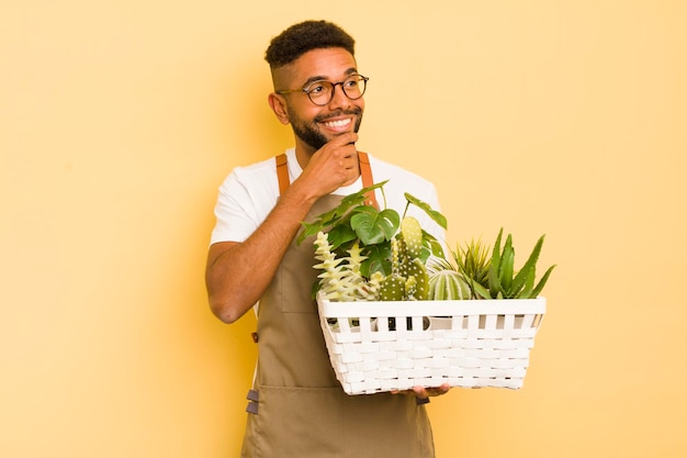 Afro fajny mężczyzna uśmiechający się ze szczęśliwym, pewnym wyrazem twarzy z ręką na podbródku ogrodnik i koncepcja roślin